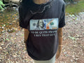 A person wearing a black graphic t-shirt featuring a printed $100 bill and the text 'TO BE QUITE FRANK I BEN THAT GUY' in a real outdoor setting with a stream and rocks in the background.