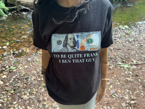 A person wearing a black graphic t-shirt featuring a printed $100 bill and the text 'TO BE QUITE FRANK I BEN THAT GUY' in a real outdoor setting with a stream and rocks in the background.