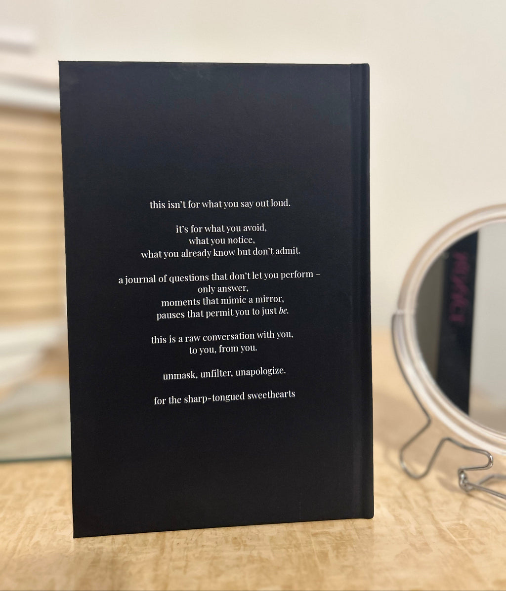 A black hardcover journal standing upright on a light wood surface, with white printed text on the cover and a blurred round mirror in the background.