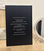 A black hardcover journal standing upright on a light wood surface, with white printed text on the cover and a blurred round mirror in the background.