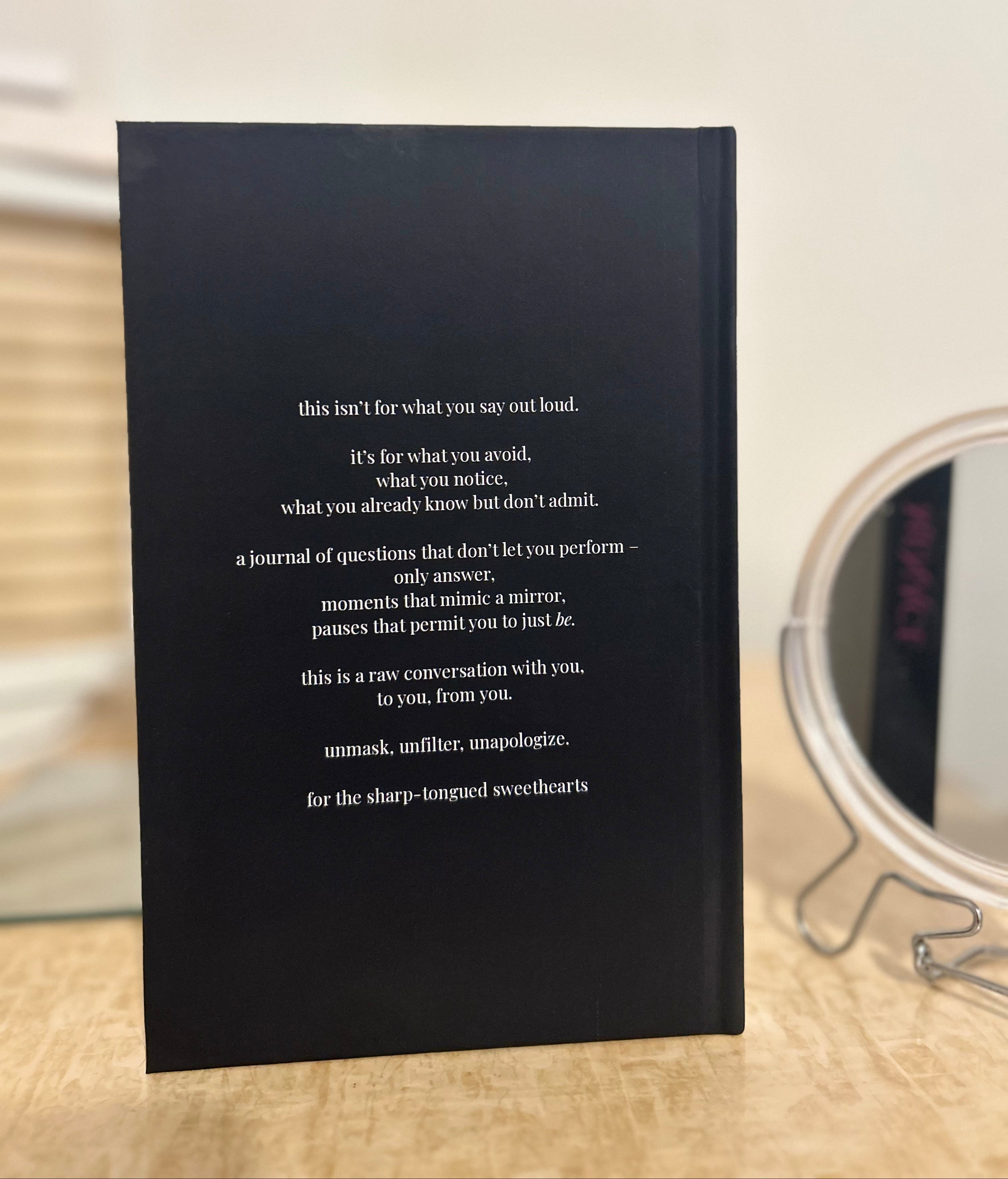 A black hardcover journal standing upright on a light wood surface, with white printed text on the cover and a blurred round mirror in the background.