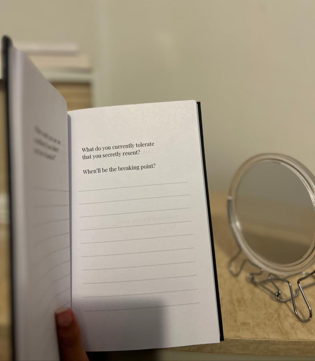 An open notebook or journal with lined pages and printed introspective prompts, held open by a hand in a home setting with a small round mirror on a table in the background.