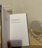 An open notebook or journal with lined pages and printed introspective prompts, held open by a hand in a home setting with a small round mirror on a table in the background.