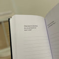 An open notebook or journal held in a hand, showing printed introspective text prompts on the pages against a softly blurred indoor background.