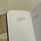 An open notebook or journal held in a hand, showing printed introspective text prompts on the pages against a softly blurred indoor background.