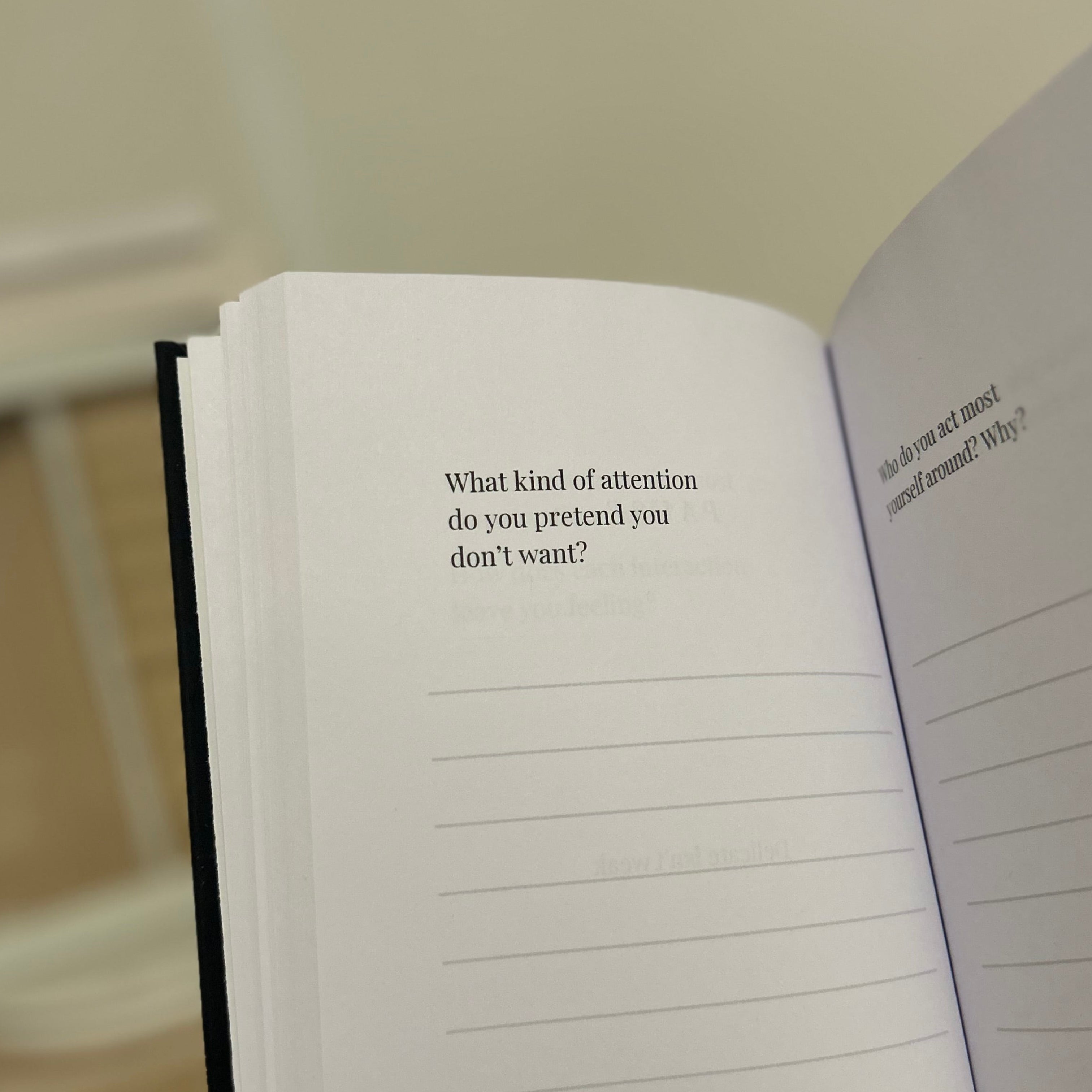 An open notebook or journal held in a hand, showing printed introspective text prompts on the pages against a softly blurred indoor background.