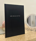 A black hardcover book or journal standing upright on a light beige surface, with the phrase 'write what you won't say' printed in small white text on the front. A small round mirror is blurred in the background.