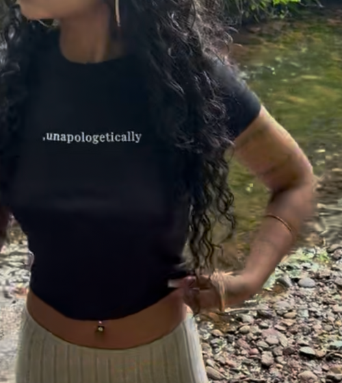 A woman standing outdoors near a rocky stream wearing a black short-sleeve crop top with white text and a long ribbed beige skirt.