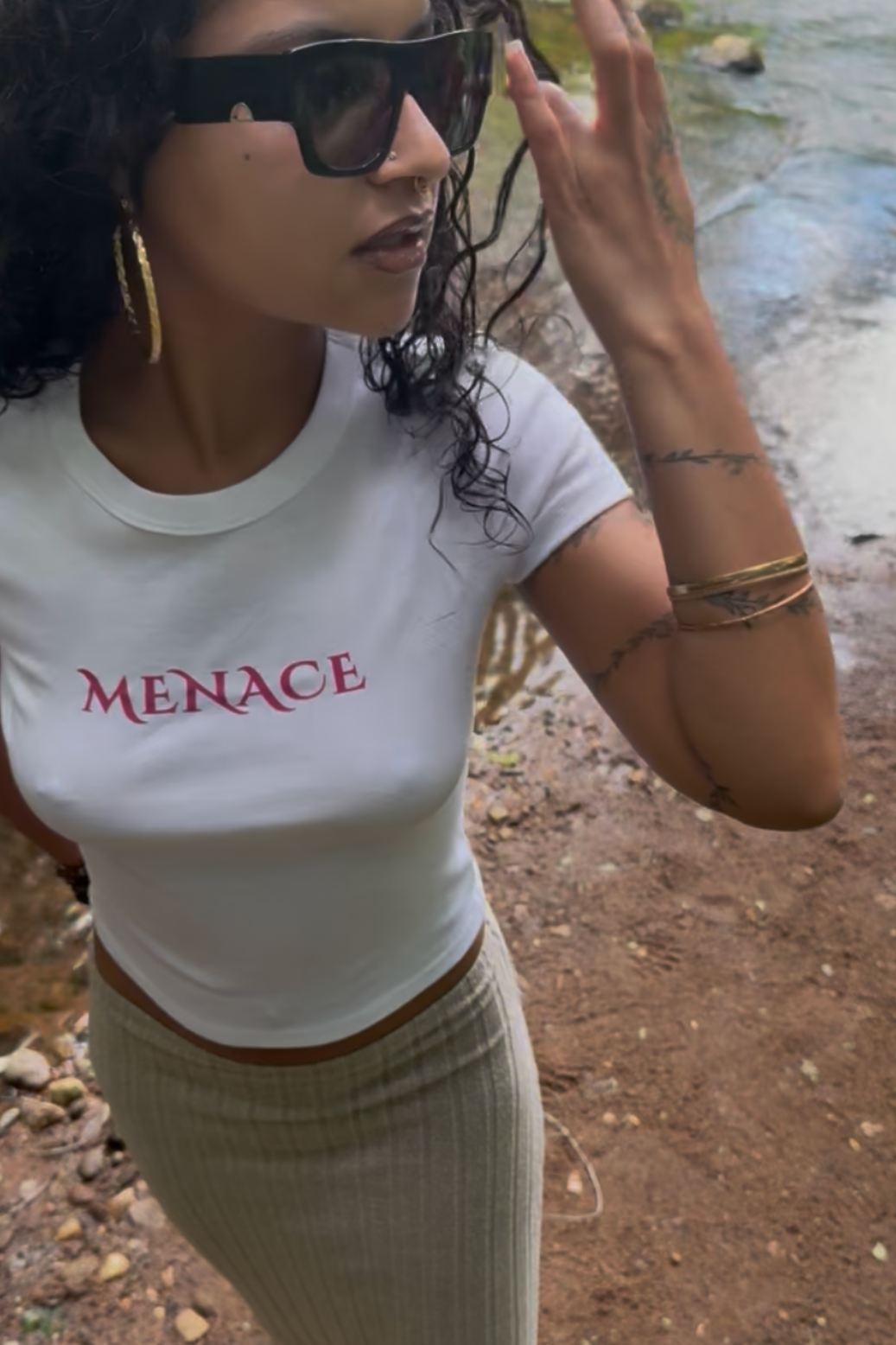 A woman wearing a white short-sleeve crop top with the word 'MENACE' printed in pink across the chest, paired with a beige ribbed maxi skirt and black sunglasses, standing outdoors near water.